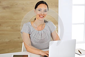 Cheerful smiling business woman working with laptop computer while sitting at the desk in modern office. Middle aged