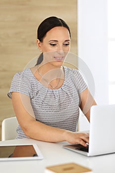 Cheerful smiling business woman working with laptop computer while sitting at the desk in modern office. Middle aged