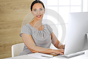 Cheerful smiling business woman working with pc computer while sitting at the desk in modern office. Middle aged female