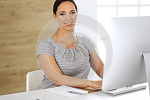 Cheerful smiling business woman working with pc computer while sitting at the desk in modern office. Middle aged female