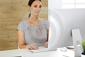 Cheerful smiling business woman working with pc computer while sitting at the desk in modern office. Middle aged female