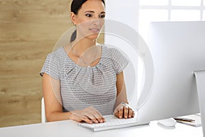 Cheerful smiling business woman working with pc computer while sitting at the desk in modern office. Middle aged female