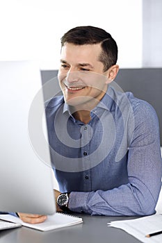 Cheerful smiling businessman working with computer in modern office. Headshot of male entrepreneur or director of a