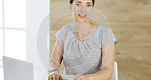 Cheerful smiling businesswoman working with laptop computer while sitting at the desk in modern office. Middle aged