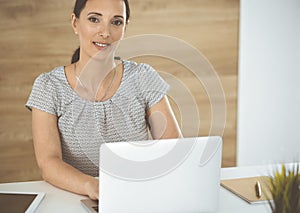 Cheerful smiling businesswoman working with laptop computer while sitting at the desk in modern office. Middle aged