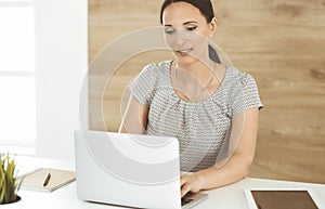 Cheerful smiling businesswoman working with laptop computer while sitting at the desk in modern office. Middle aged