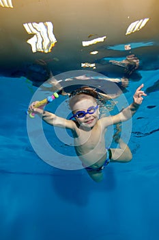 A cheerful, smiling child, a little boy, is swimming underwater in the pool. He holds sports toys in his hands and looks