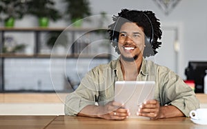 Cheerful smiling curly young black guy in wireless headphones at tablet, listen music and looking at empty space