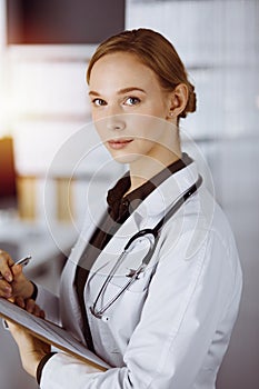 Cheerful smiling female doctor using clipboard in clinic. Portrait of friendly physician woman at work. Medical service