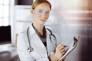Cheerful smiling female doctor using clipboard in clinic. Portrait of friendly physician woman at work. Medical service