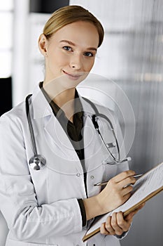 Cheerful smiling female doctor using clipboard in clinic. Portrait of friendly physician woman at work. Perfect medical