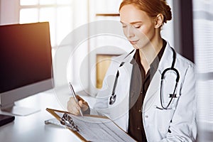 Cheerful smiling female doctor using clipboard in clinic. Portrait of friendly physician woman at work place. Medical