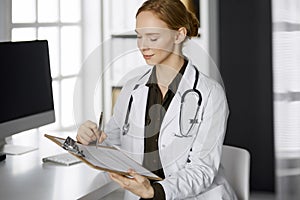 Cheerful smiling female doctor using clipboard in clinic. Portrait of friendly physician woman at work place. Perfect