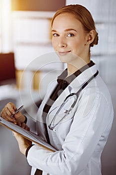 Cheerful smiling female doctor using clipboard in sunny clinic. Portrait of friendly physician woman at work. Medicine