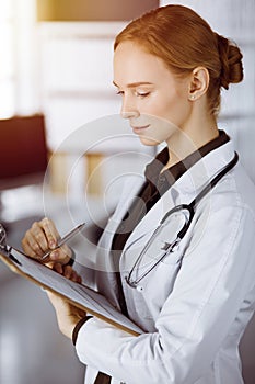 Cheerful smiling female doctor using clipboard in sunny clinic. Portrait of friendly physician woman at work. Medicine