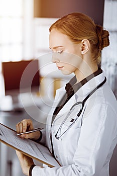 Cheerful smiling female doctor using clipboard in sunny clinic. Portrait of friendly physician woman at work. Medicine