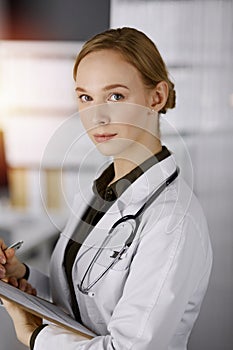 Cheerful smiling female doctor using clipboard in sunny clinic. Portrait of friendly physician woman at work. Perfect