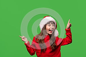 A cheerful smiling girl in a Santa hat shows with her index fingers discounts, promotions, gifts. Emotional portrait