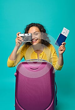 Cheerful smiling Hispanic woman holding a passport with ticket and boarding pass and a vintage camera, photographing, smiling