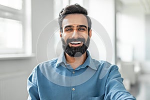 Cheerful smiling indian man wearing shirt sitting at office, closeup photo of happy businessman at workplace