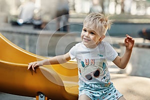 A cheerful, smiling, likeable little boy on the playground on a yellow slider. The child's hair is electrified and bristling