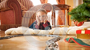 Cheerful smiling little boy looking on circle railroad under Christmas tree at houe living room. Child receiving