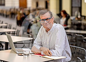 Cheerful smiling old man working on computer while having coffee in terrace coffee shop city outdoors in seniors using modern