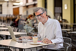 Cheerful smiling old man working on computer while having coffee in terrace coffee shop city outdoors in seniors using modern