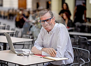 Cheerful smiling old man working on computer while having coffee in terrace coffee shop city outdoors in seniors using modern