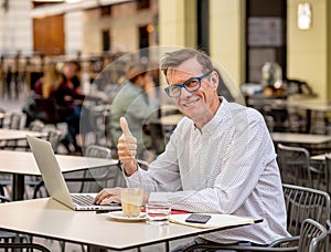 Cheerful smiling old man working on computer while having coffee in terrace coffee shop city outdoors in seniors using modern