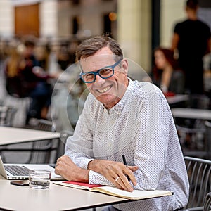 Cheerful smiling old man working on computer while having coffee in terrace coffee shop city outdoors in seniors using modern