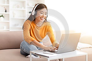 Cheerful young asian woman using laptop and headphones at home