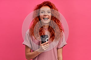 Cheerful smiling red-haired pretty girl holds takeaway coffee on pink background
