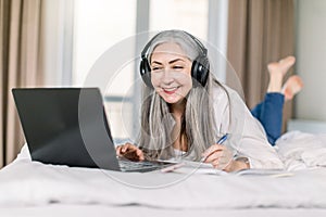 Cheerful smiling retired woman in headphones lying on bed at home, using laptop and making notes in her notepad, working