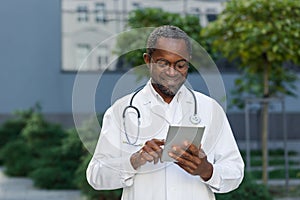 Cheerful and smiling senior doctor outside clinic using tablet computer, african american man wearing glasses browsing