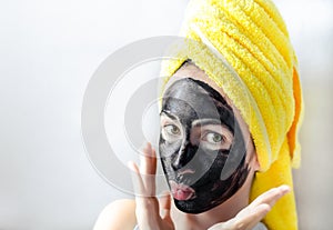 Cheerful smiling teen girl portrait with black facial clay mask and yellow towel on head over white background. Spa