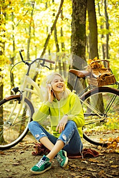 Cheerful smiling teenage girl sitting on fall autumn leaves and listening to music. Blonde stylish woman with bicycle