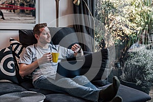 Cheerful smiling young adult man comfortably sitting at home on large sofa using laptop computer drinking coffe or tea spending