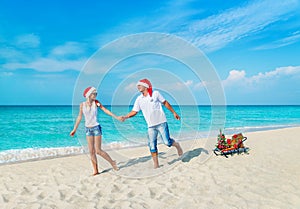 Cheerful smiling young couple in red Santa hats walking at tropical ocean sandy beach with sleds decorated fir-tree and golden fan