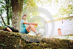Cheerful smiling young female backpacker sitting on the old stone castle fance and enjoying a rest time on the Way of Camino de Sa
