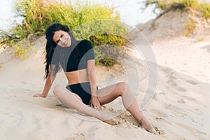 A cheerful smiling young girl came to rest on the sea, enjoys the sun, sits on the sand and smiles, having a slim figure