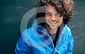 Cheerful smiling young man with curly hair sitting on stairs of a building with backpack, showing thumb up. Happy male resting in