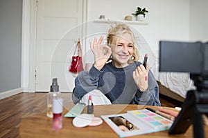 Cheerful, smiling young woman recording a vlog, showing okay, ok gesture and favorite lipstick, recommending beauty