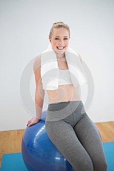 Cheerful sporty blonde with towel around her neck sitting on exercise ball