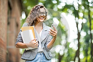 A cheerful student with multi-colored notebooks and a bag is walking around the campus and talking on the phone