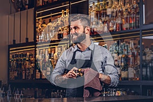 Cheerful stylish brutal barman is cleaning the glass with a cloth at bar counter background.