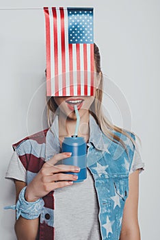 Cheerful stylish girl in american patriotic outfit drinking soda from can, with american flag in front of her face