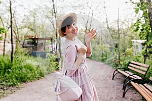 Cheerful stylish girl goes to the wooden bench to rest after long walk in sunny day. Portrait of slim smiling young