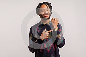 Cheerful successful african man with dreadlocks in stylish sweatshirt pointing finger on his watch with toothy smile on face,