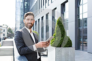 Cheerful and successful businessman works outdoors near the office holds a tablet, smiles reads good news